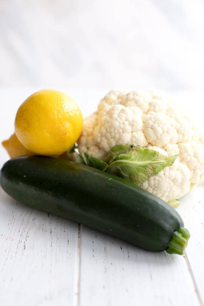 A head of cauliflower, a medium zucchini, and two lemons on a white table.