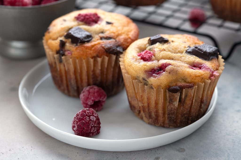 Two keto raspberry chocolate chip muffins on a white plate with frozen raspberries.