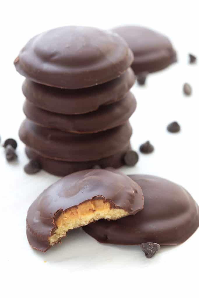 Two keto tagalong cookies in front of a stack of more cookies, with chocolate chips strewn around.