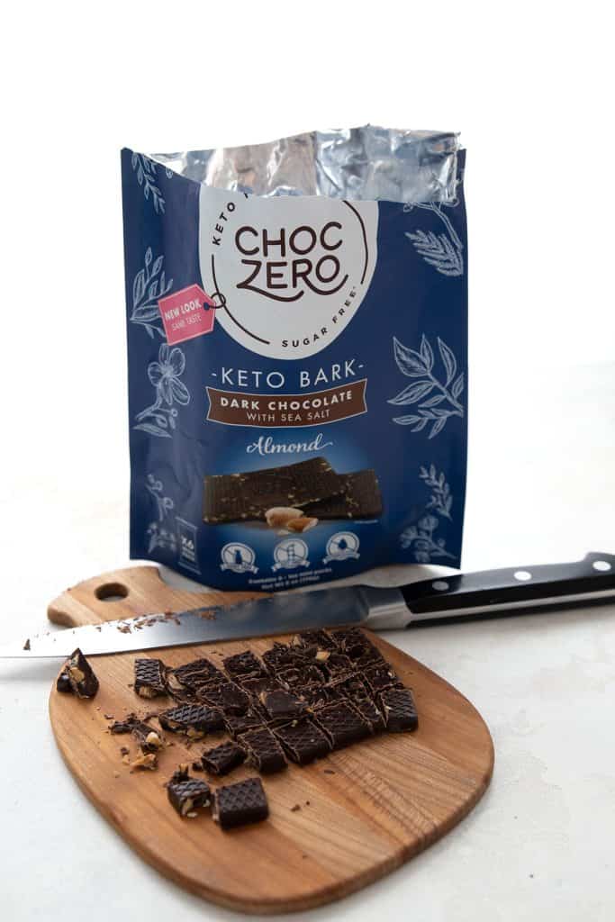 Keto chocolate chunks on a wooden cutting board with a knife, and a bag of keto chocolate bark in the background.