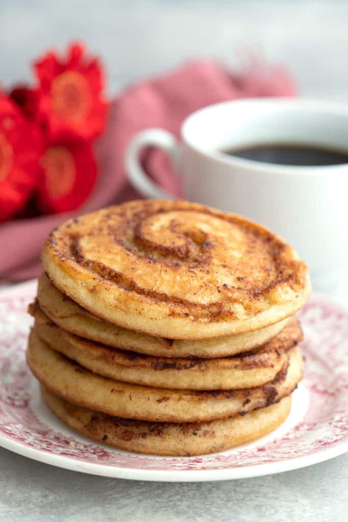 A stack of keto cinnamon roll pancakes with a cup of coffee and red flowers in the background.
