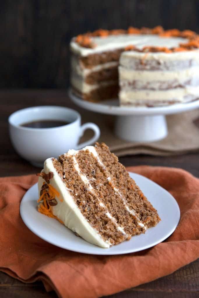 A slice of keto carrot cake lying on a white plate with the rest of the cake and a cup of coffee in the background.