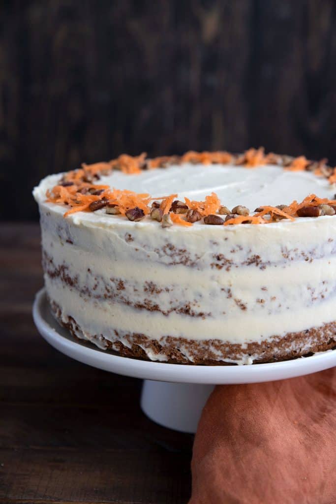 Keto Carrot Cake on a white cake platter, decorated with chopped pecans and shredded carrot.