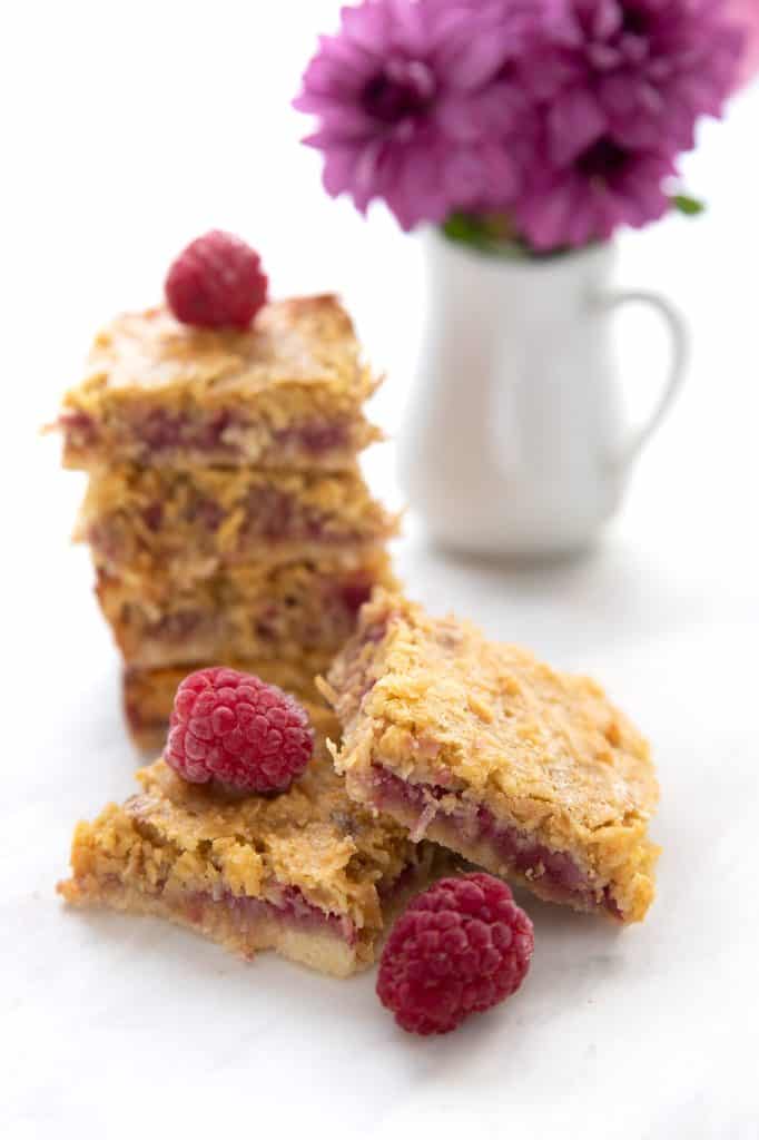 Two raspberry coconut bars in front of a stack of more bars and a vase with flowers.