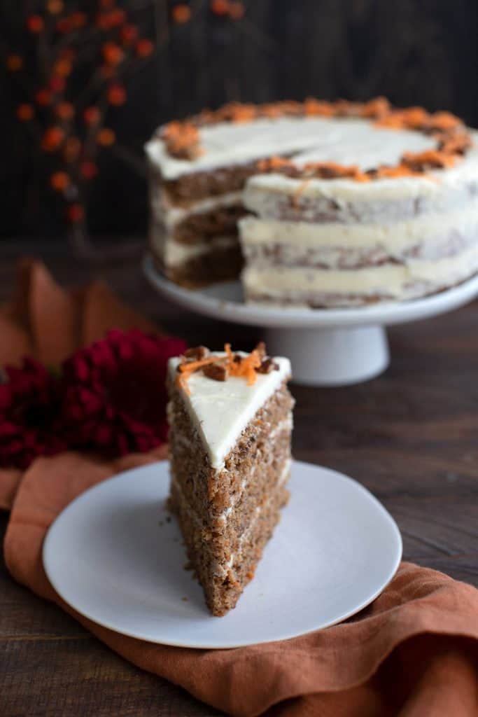 A tall slice of keto carrot layer cake on a white plate with the rest of the cake in the background.