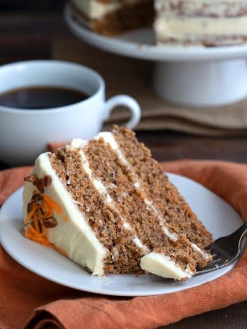 A slice of keto carrot cake on a white plate with a forkful taken out of it and a cup of coffee in the background.