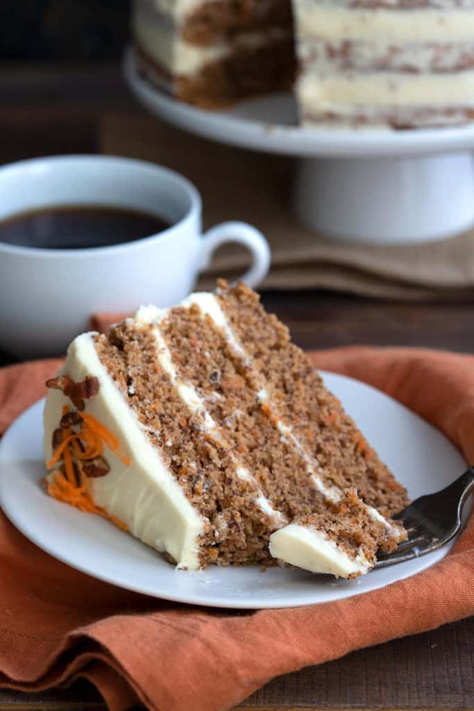 A slice of keto carrot cake on a white plate with a forkful taken out of it and a cup of coffee in the background.