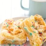 Two Keto Sugar Cookie Cheesecake Bars on a small white plate with a small cup of coffee in the background.