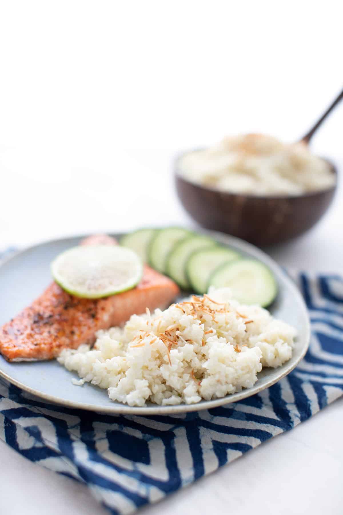 A blue plate filled with coconut cauliflower rice, salmon, and cucumber over a blue patterned napkin.