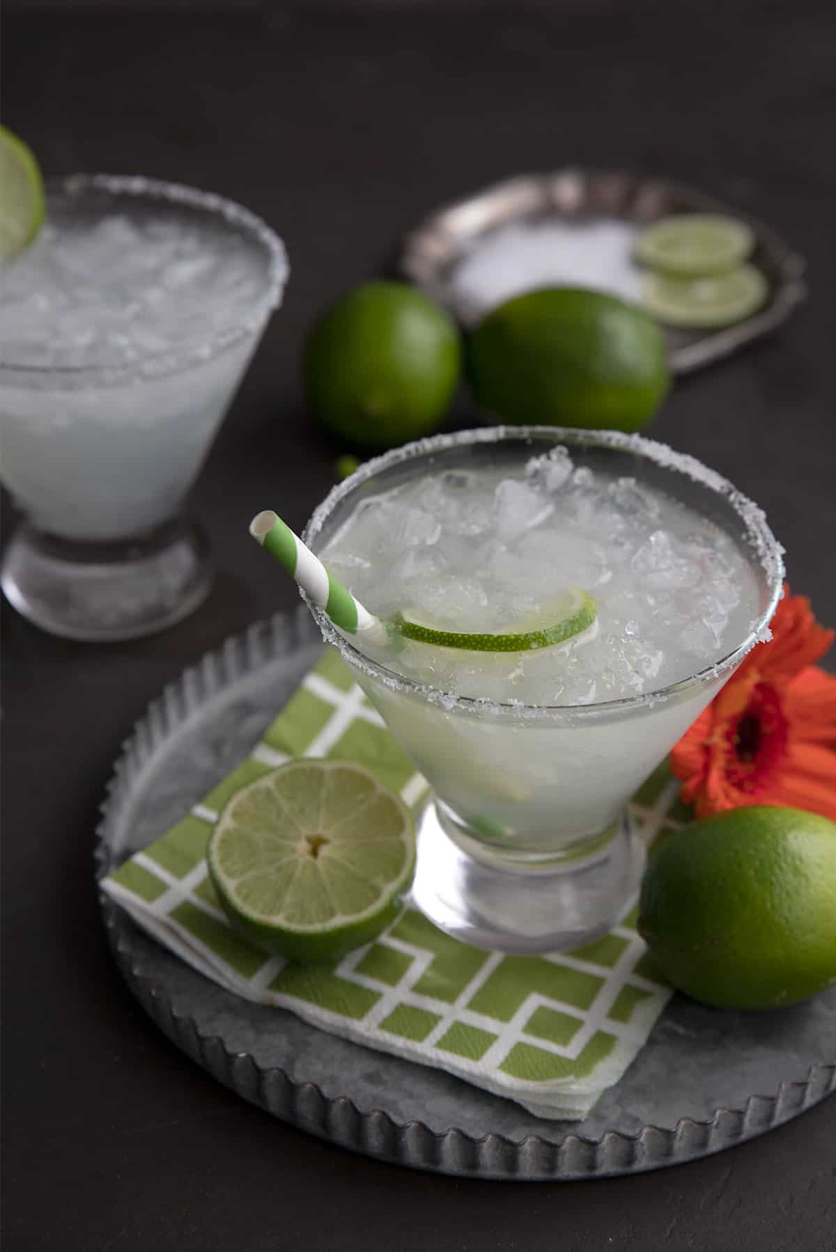 Two keto margaritas in cocktail glasses on a pewter tray, with sliced limes and a dish of salt.