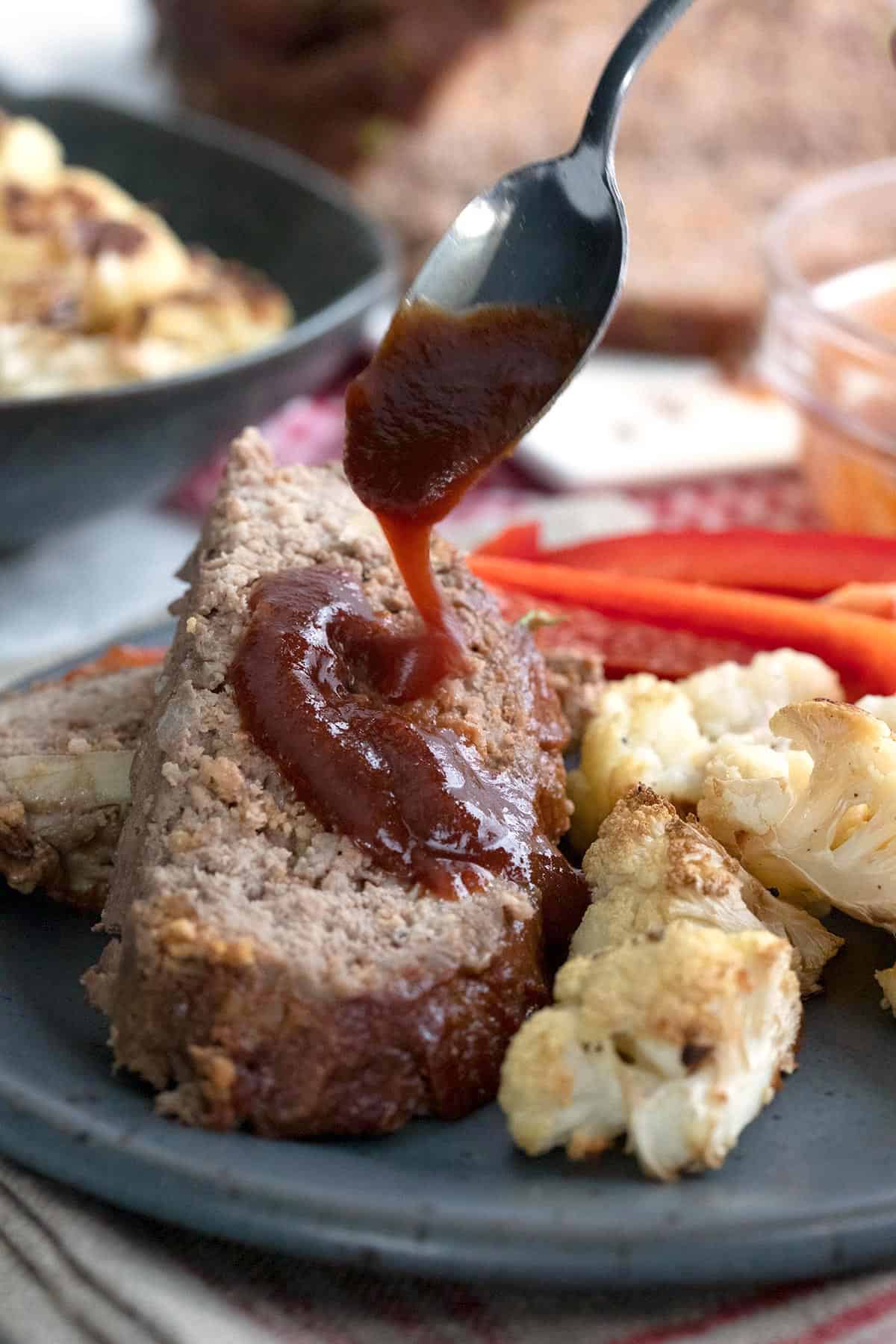 A spoon drizzling sugar free bbq sauce over a slice of keto meatloaf.