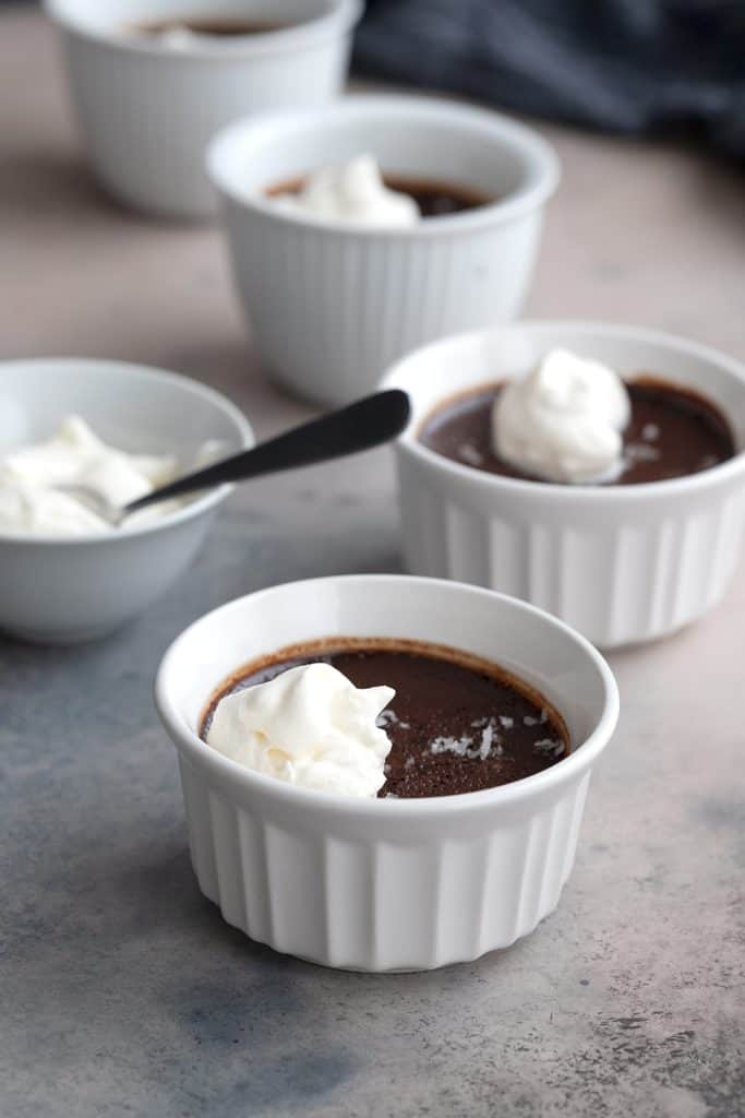 Four ramekins filled with sugar-free pot de creme on a grey table, with a bowl of whipped cream.