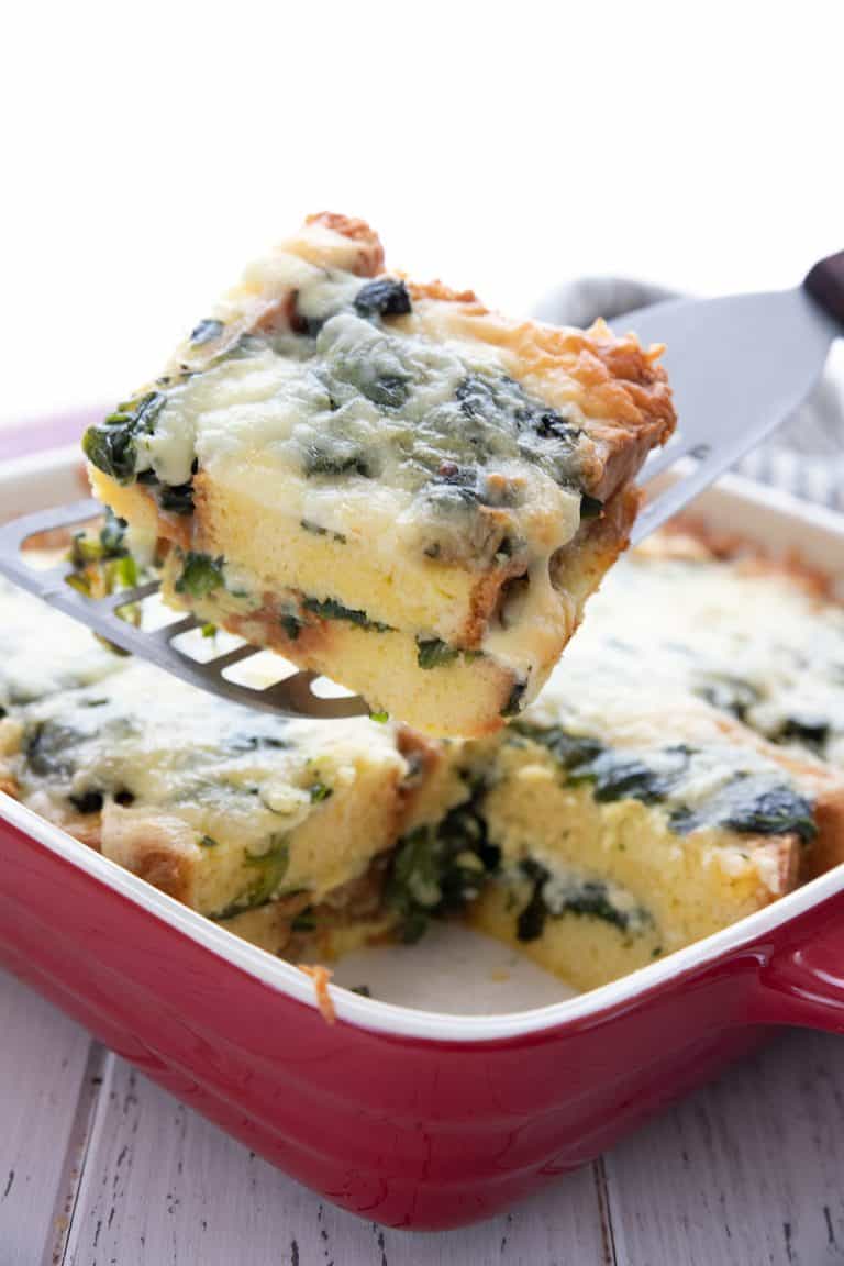 A metal spatula lifting a slice of keto egg strata out of a red baking dish.