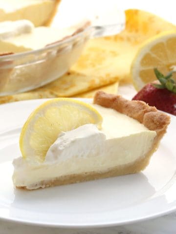 A slice of keto lemon pie on a white plate with a slice of lemon top. The remaining pie is in the background.