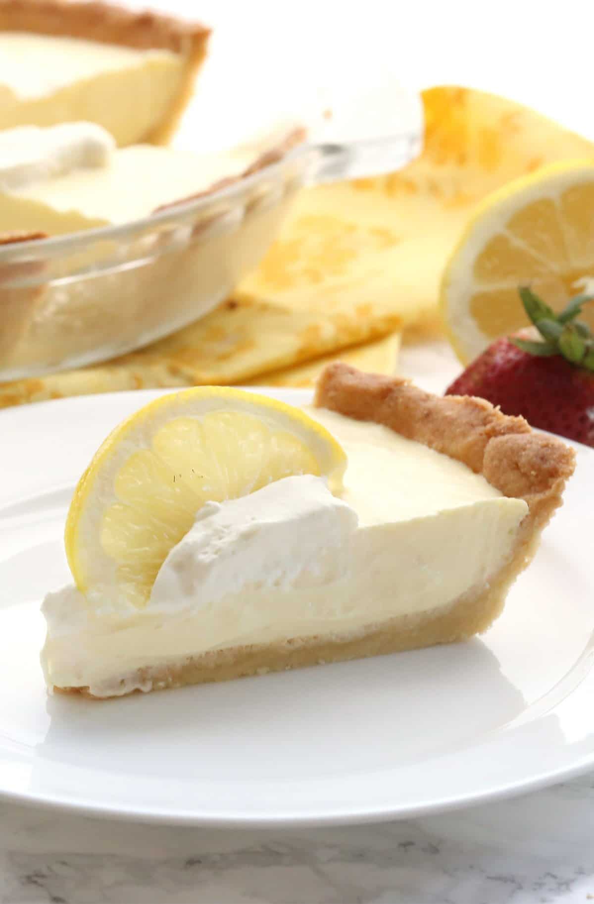 A slice of keto lemon pie on a white plate with a slice of lemon top. The remaining pie is in the background.