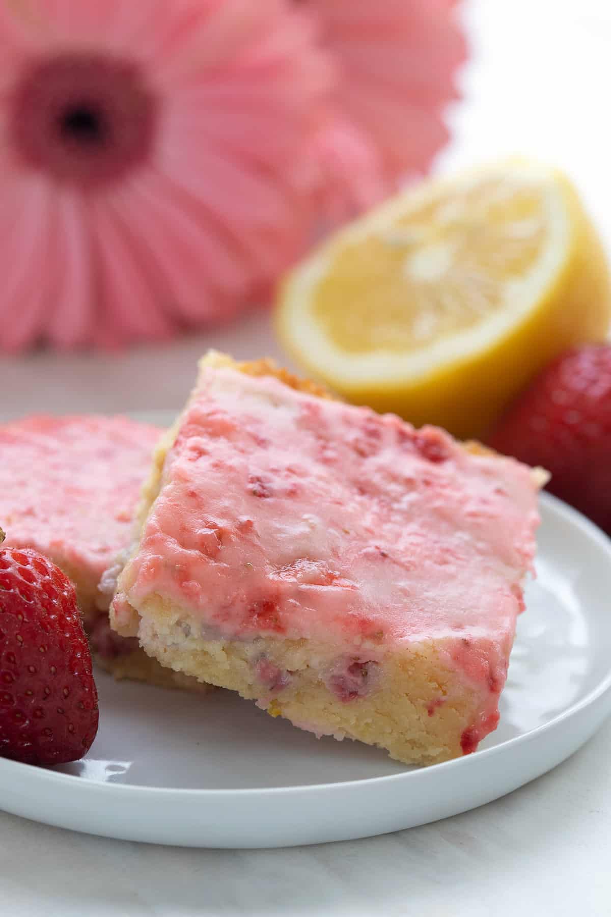 Two keto strawberry lemon blondies on a white plate with flowers, lemons, and strawberries in the background.