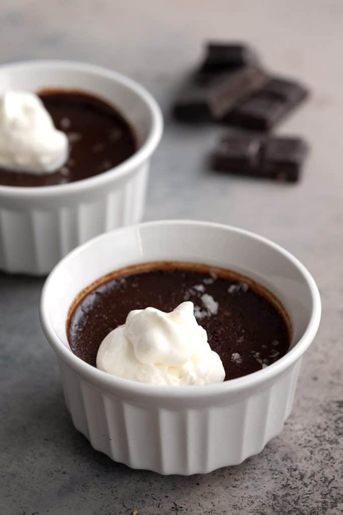 Two keto pots de creme in white ramekins with whipped cream on top and squares of chocolate in the background.