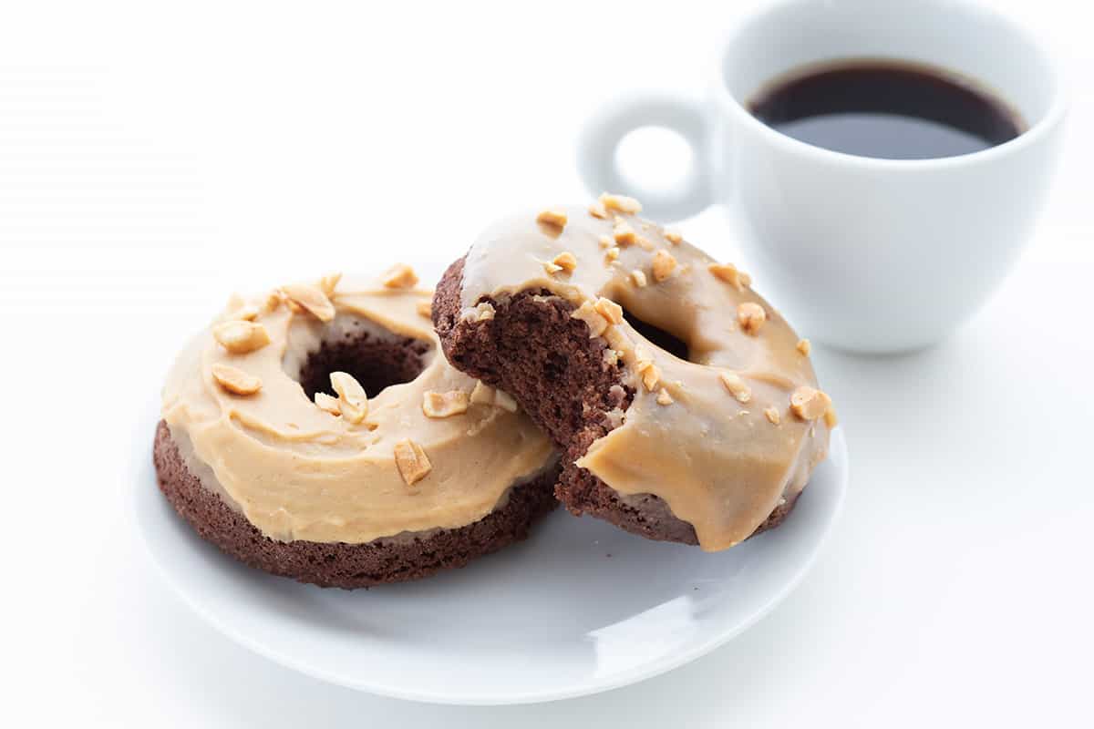 Two Keto Chocolate Peanut Butter Donuts on a white plate beside a cup of coffee.