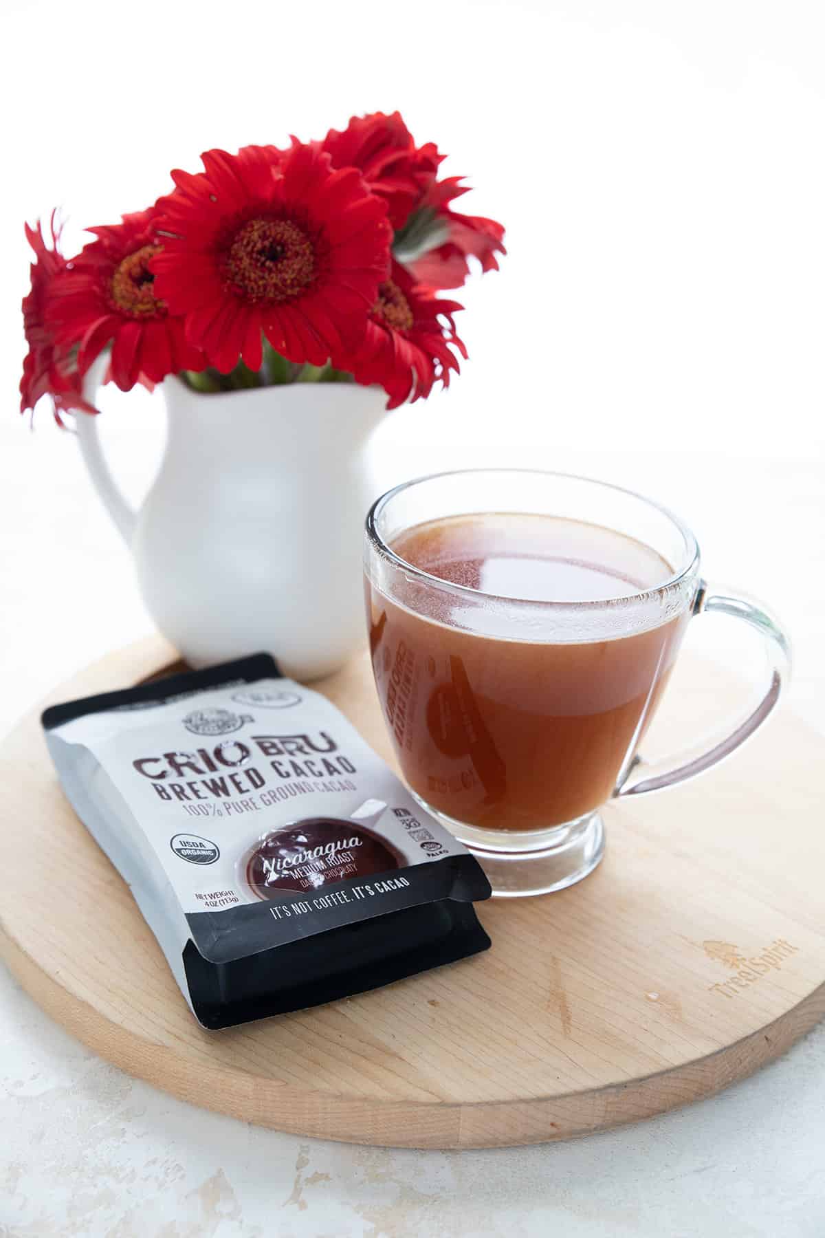 A cup of brewed cacao on a wooden platter, with red flowers and a bag of Crio Bru.