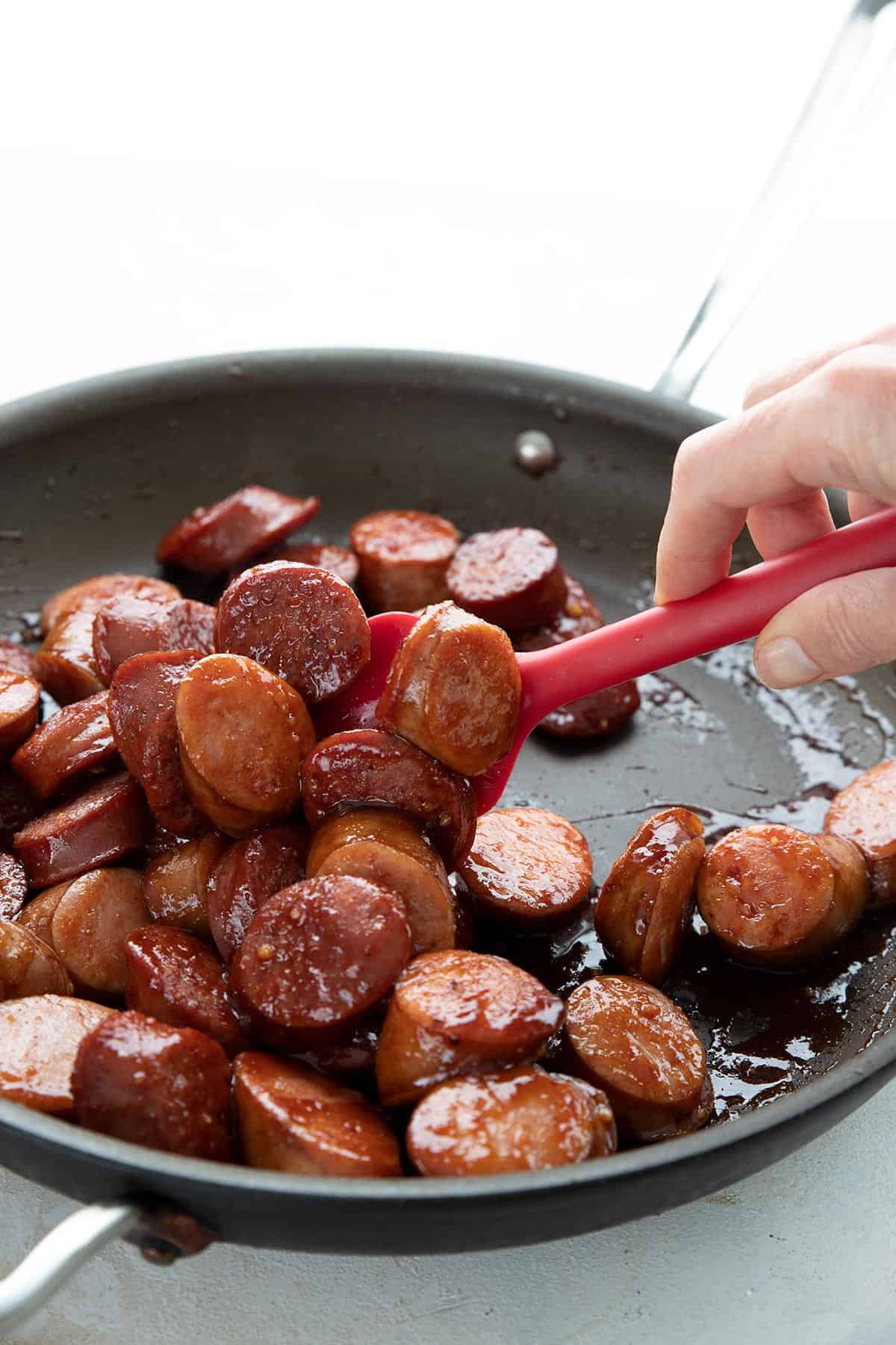 A red spatula stirring a pan filled with sweet and spicy kielbasa bites.