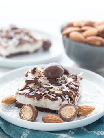 A keto no bake cheesecake bar on a white plate, over a teal napkin, with sugar-free chocolate covered almonds around it.