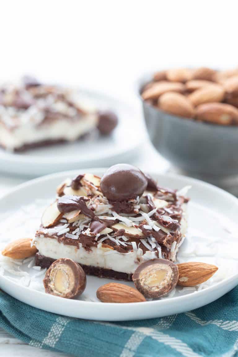 A keto no bake cheesecake bar on a white plate, over a teal napkin, with sugar-free chocolate covered almonds around it.