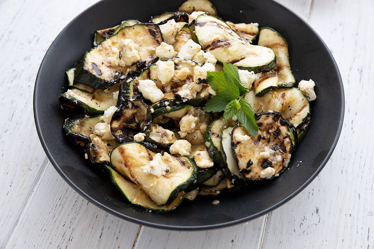 Close up shot of grilled zucchini in a black bowl, topped with feta and fresh mint.