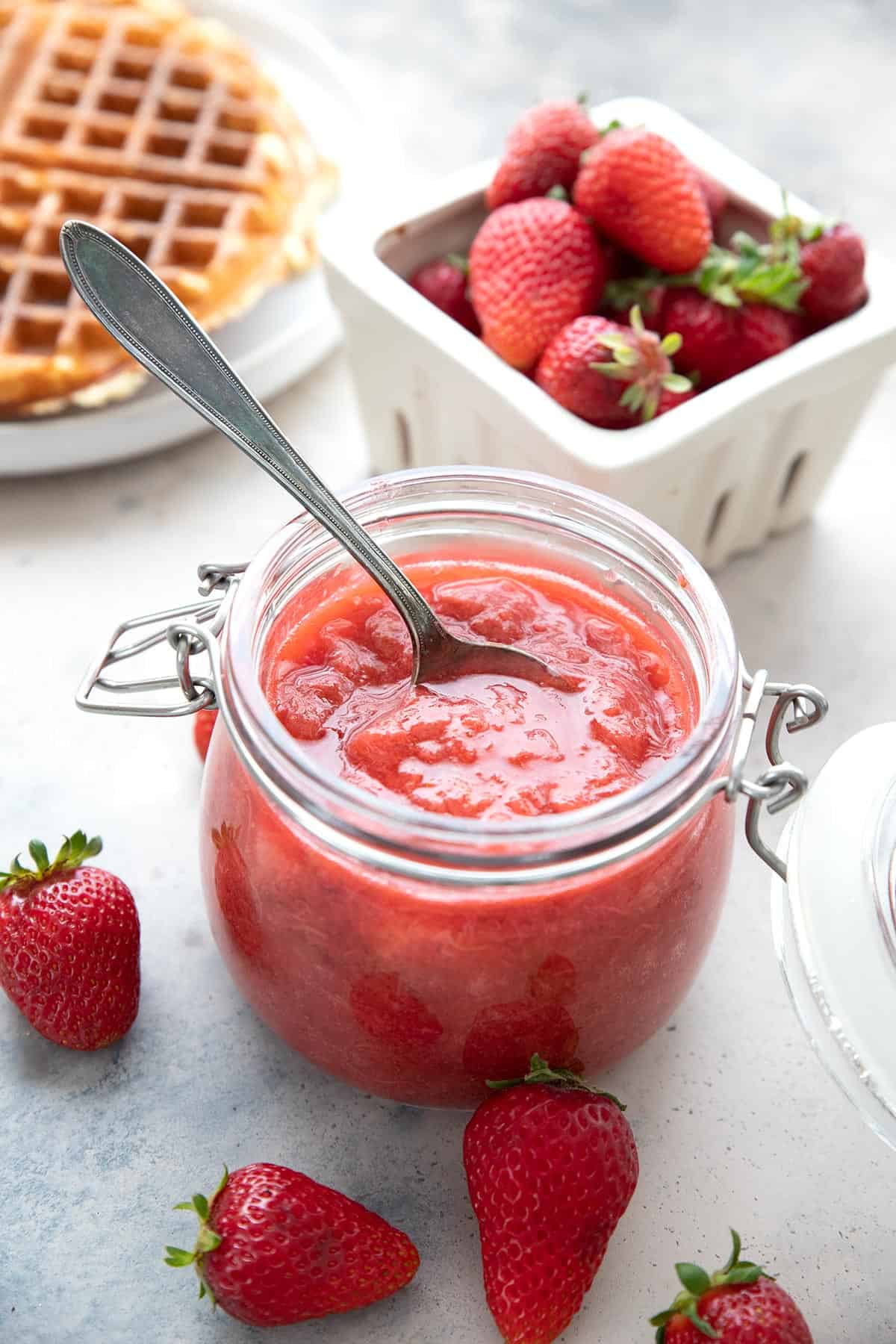 Strawberry rhubarb sauce in a jar, with a pint of strawberries and some waffles in the background.