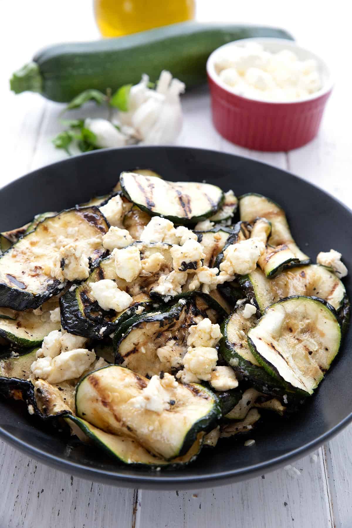 Grilled zucchini in a large black bowl with a whole zucchini, olive oil, and feta in the background.