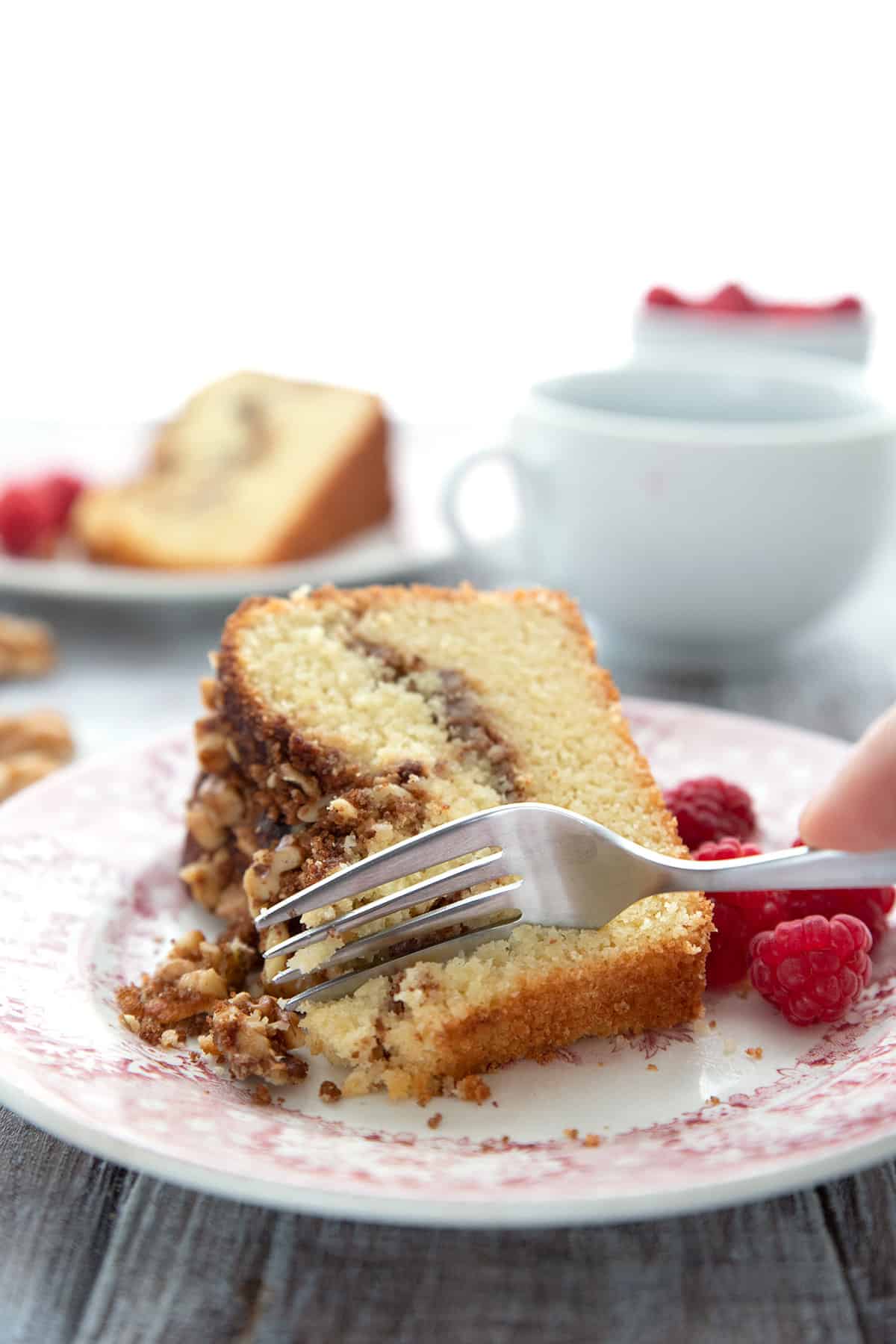 A fork digging into a slice of keto coffee cake on a red patterned plate, with raspberries on the side.