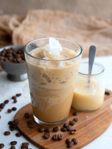 A glass of iced coffee and a jar of sweetened condensed milk on a small wooden cutting board.