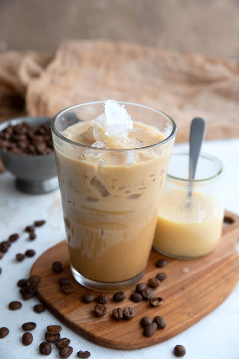 A glass of iced coffee and a jar of sweetened condensed milk on a small wooden cutting board.