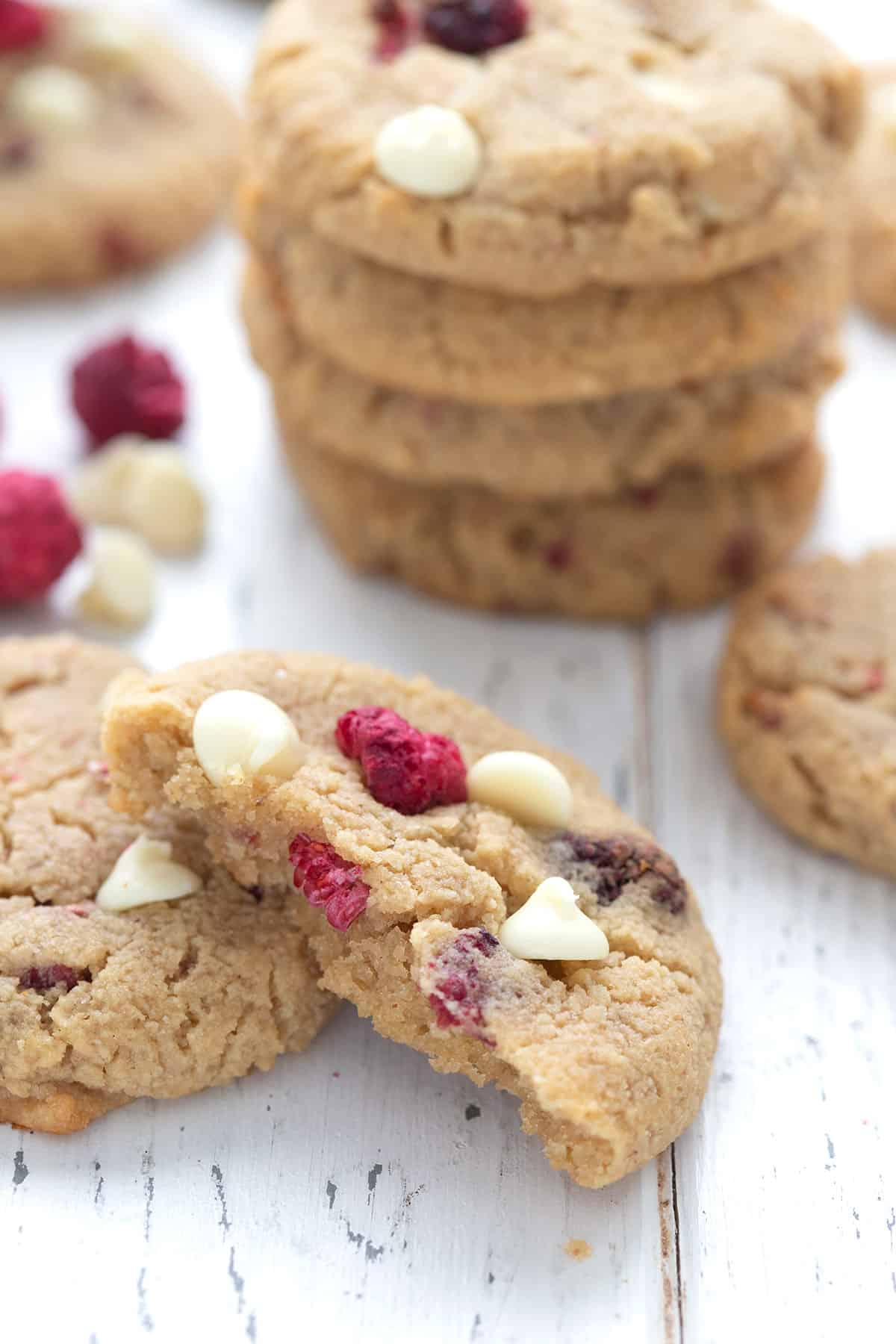A keto raspberry cheesecake cookie broken open in front of a stack of more cookies, on a white table.