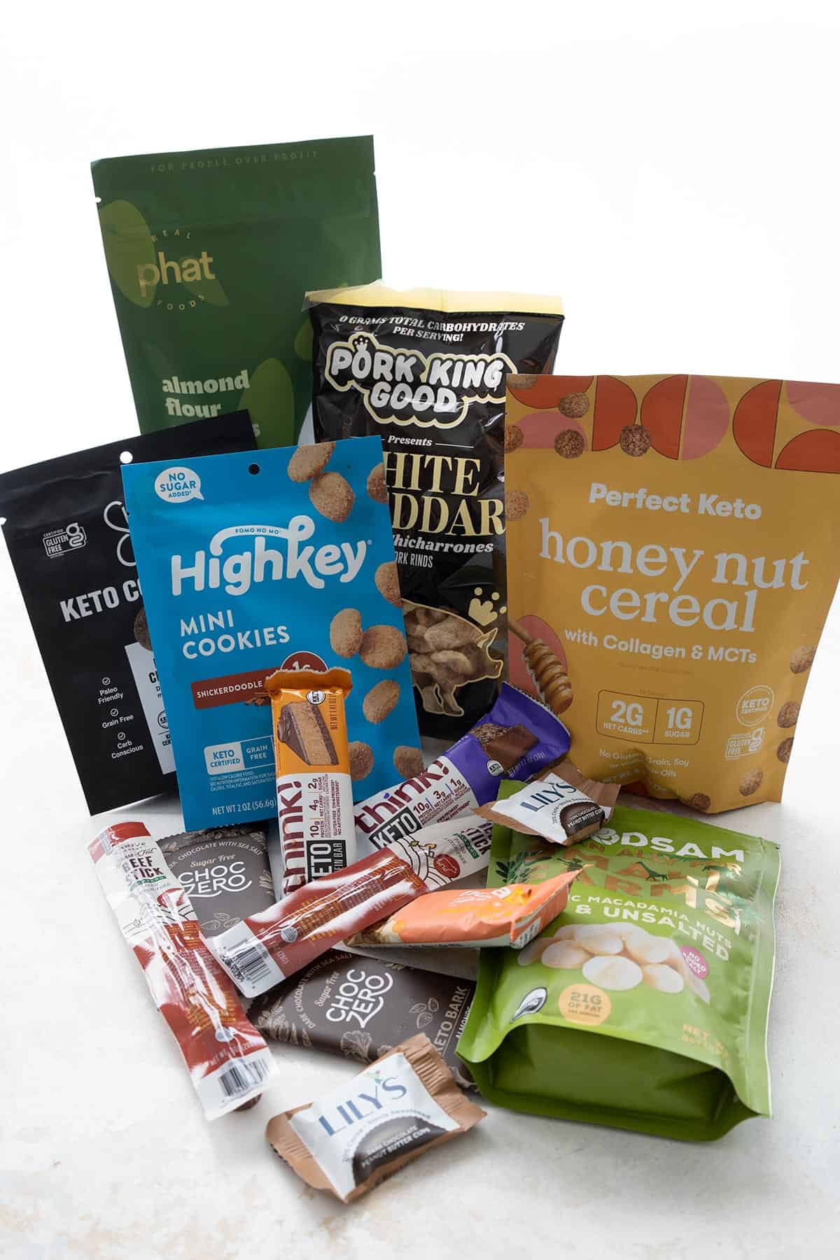 A collection of keto snacks you can by on a white table, including meat sticks, energy bars, cookies, and cereal.