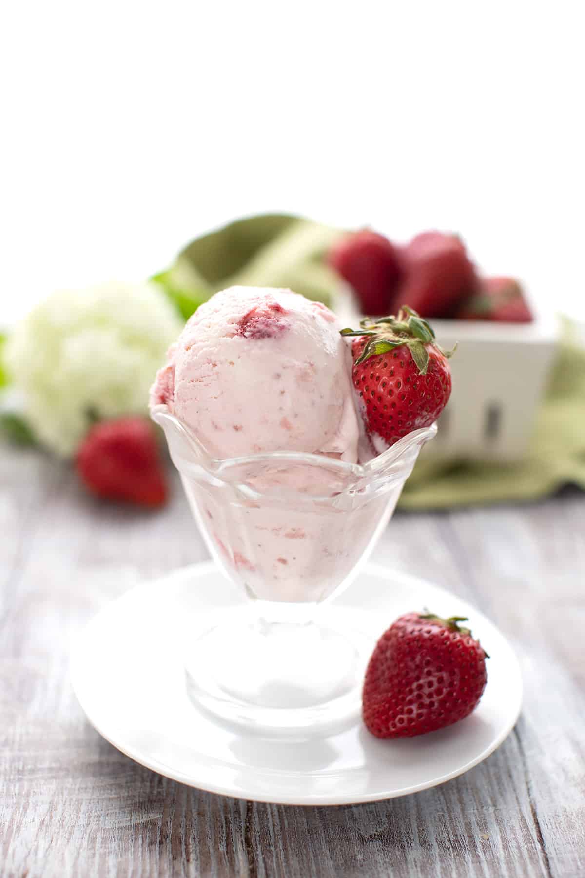 Keto strawberry ice cream in a glass ice cream cup with a fresh berry stuck in the side and another just below on the plate.