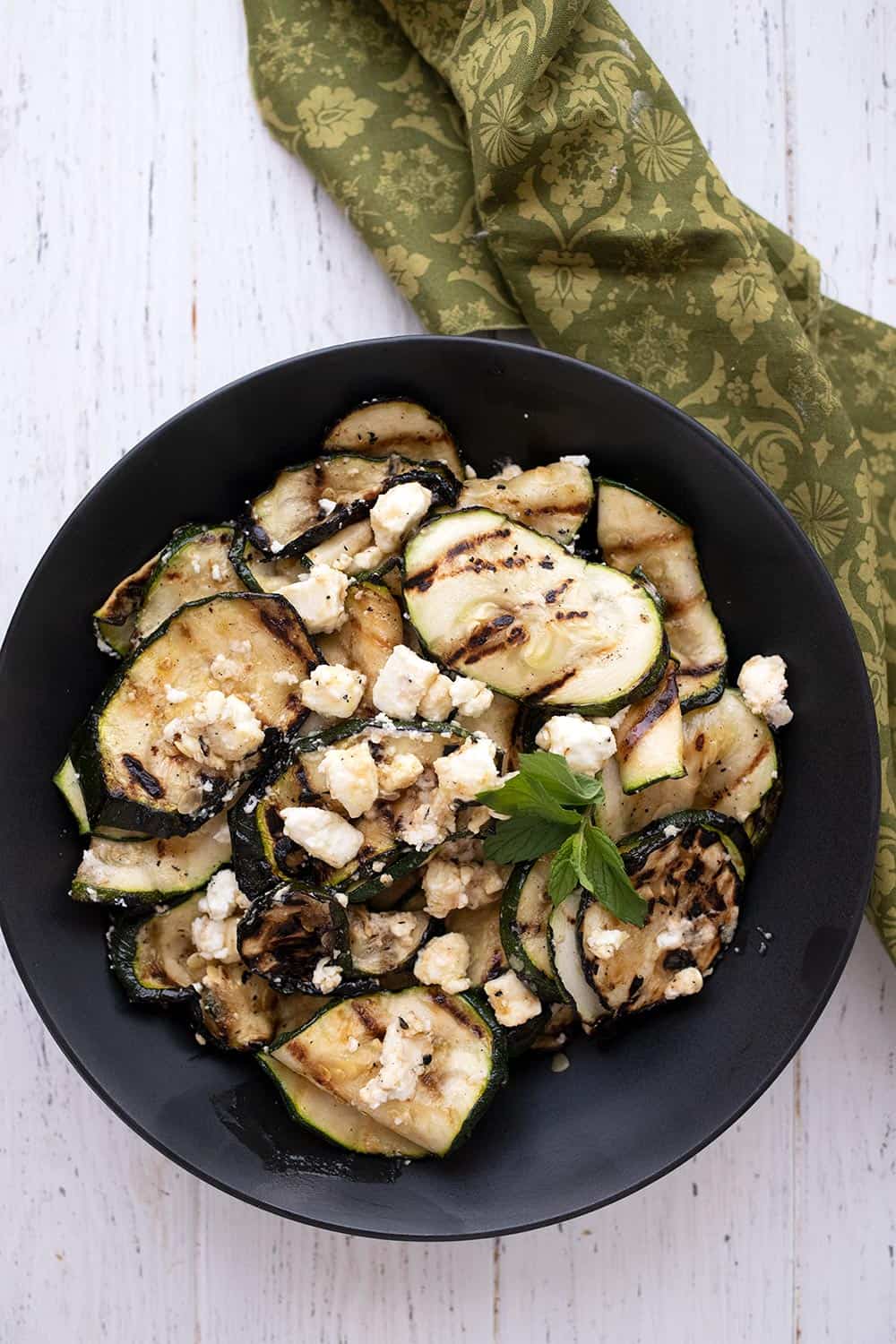 Top down image of grilled zucchini in a black bowl with a green patterned napkin.