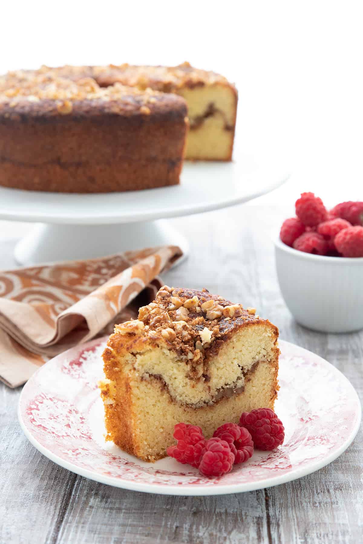 A slice of keto coffee cake on a plate, in front of the rest of the cake on a cake stand. A bowl of raspberries in the background.