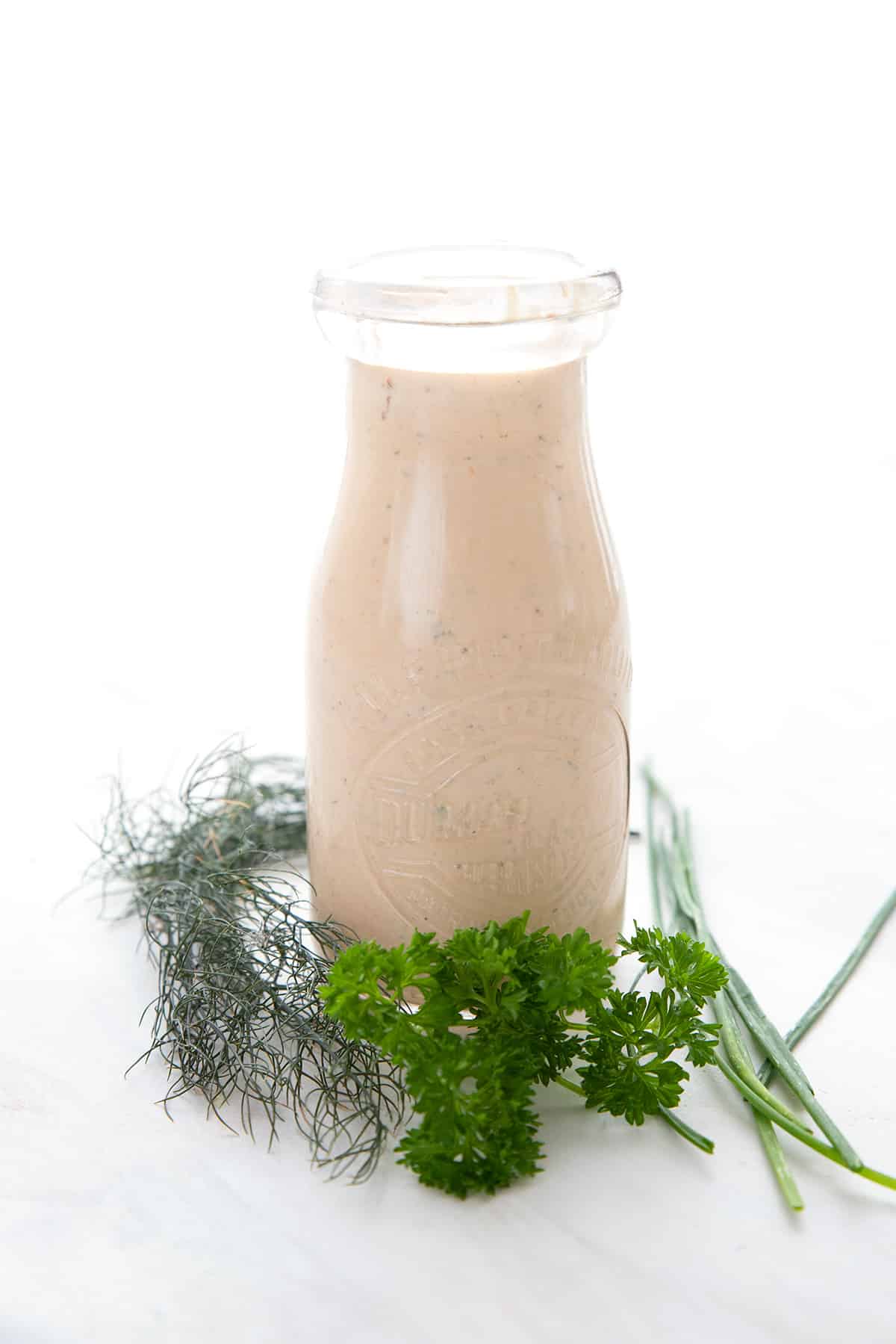 A glass bottle filled with chipotle ranch dressing surrounded by fresh herbs.