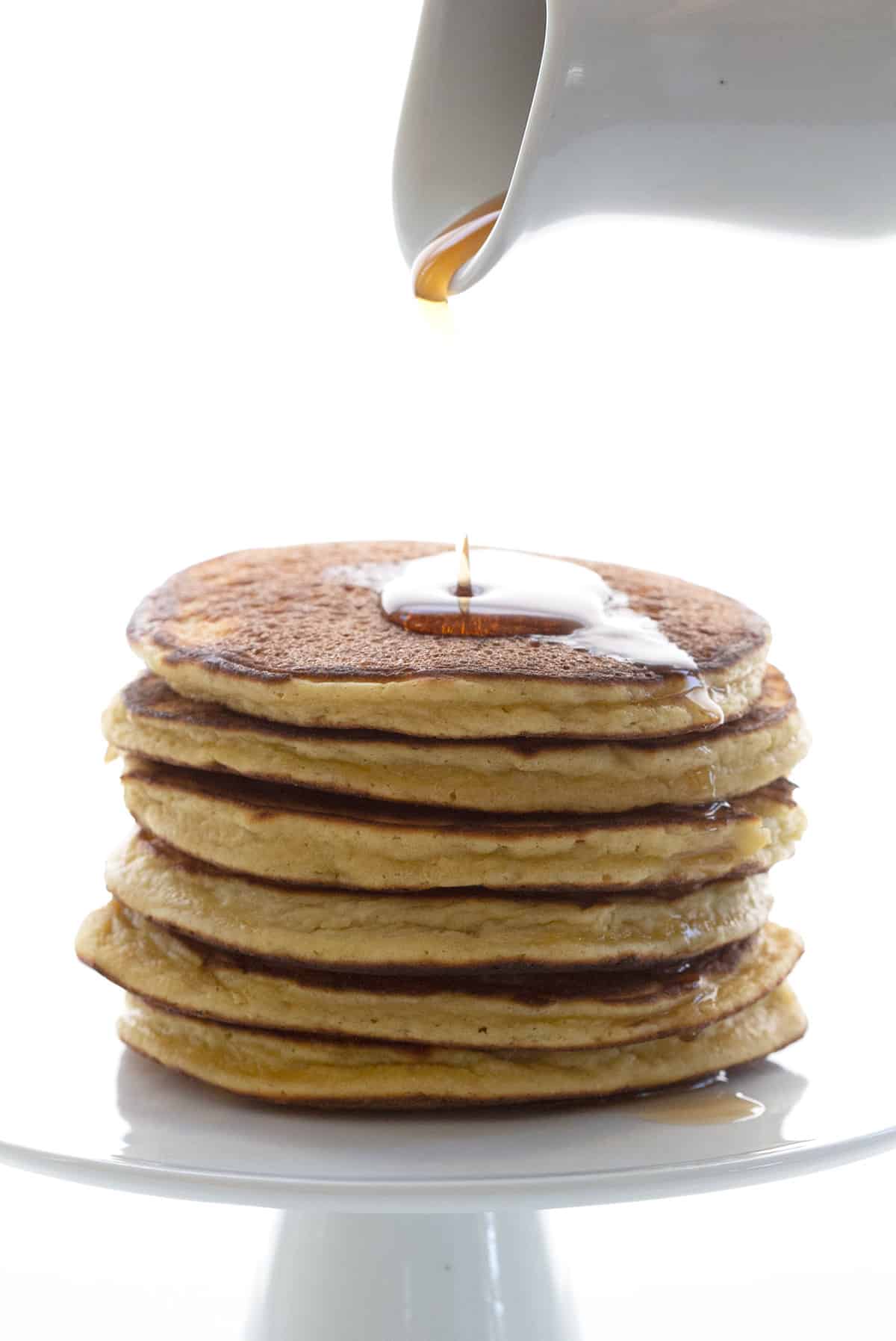 A stack of coconut flour pancakes on a cake stand with syrup being poured over top.