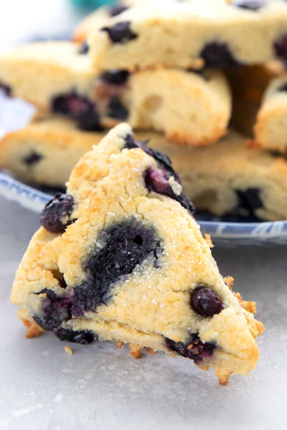 A blueberry keto scones in front of a plate of more scones on a gray table.
