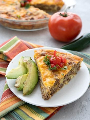 A slice of taco pie on a white plate with slices of avocado. A tomato and some jalapeno in the background.