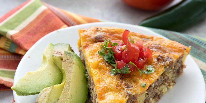 A slice of taco pie on a white plate with slices of avocado. A tomato and some jalapeno in the background.