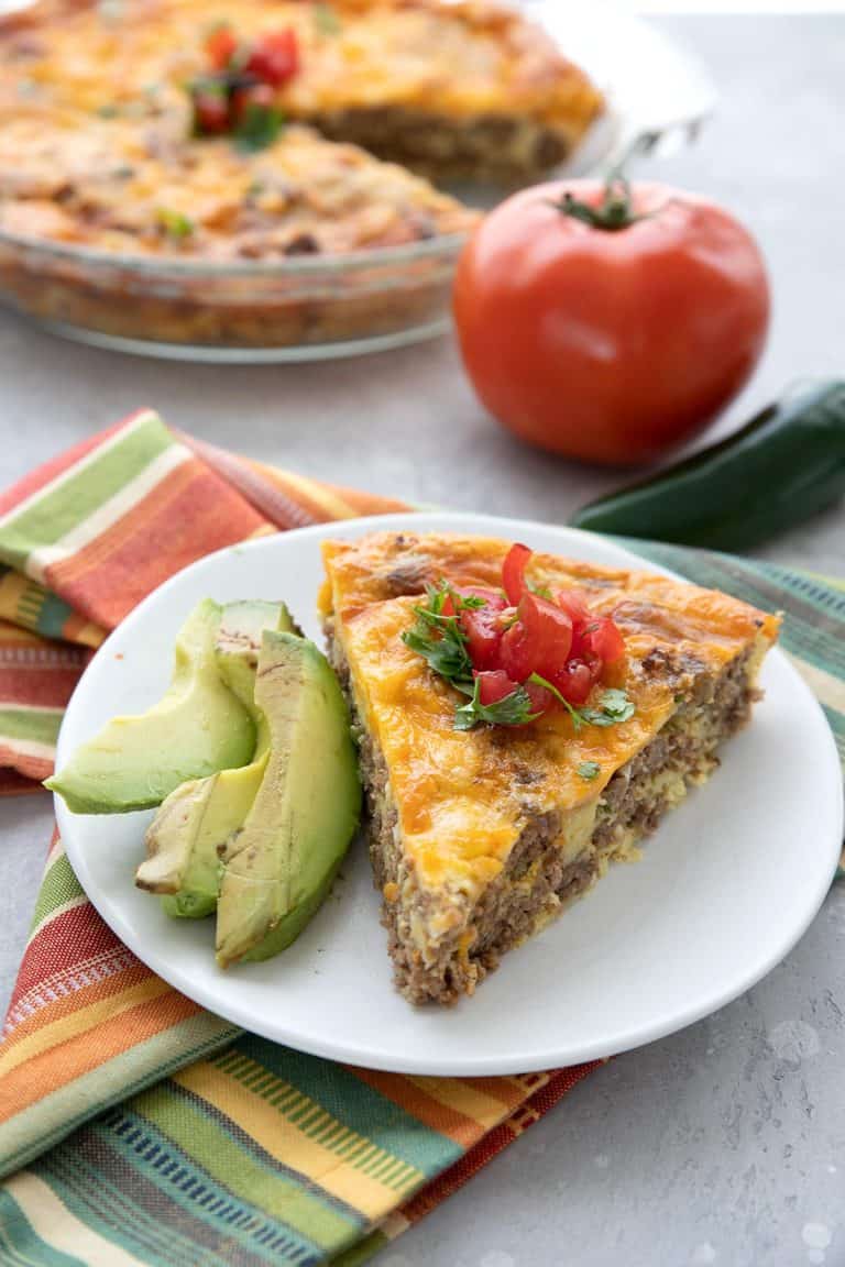 A slice of taco pie on a white plate with slices of avocado. A tomato and some jalapeno in the background.