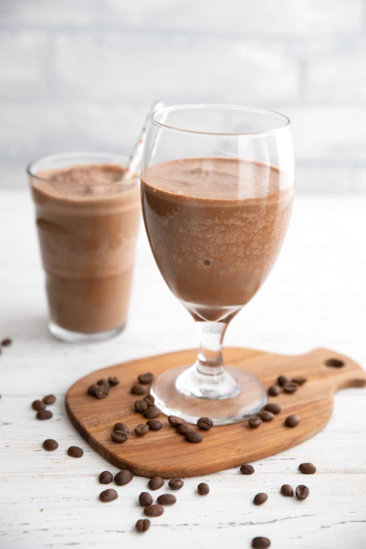 Two coffee protein shakes in glasses on a white wooden table with coffee beans strewn around. 