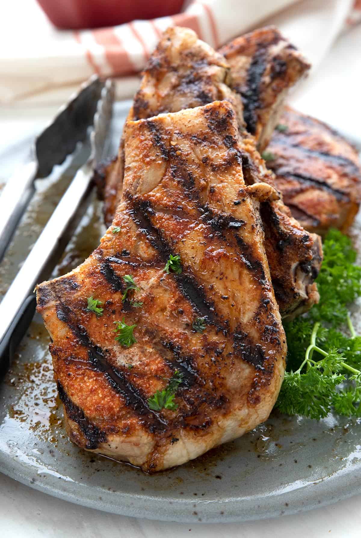 Grilled pork chops on a gray plate with sprigs of parsley and a pair of grilling tongs.