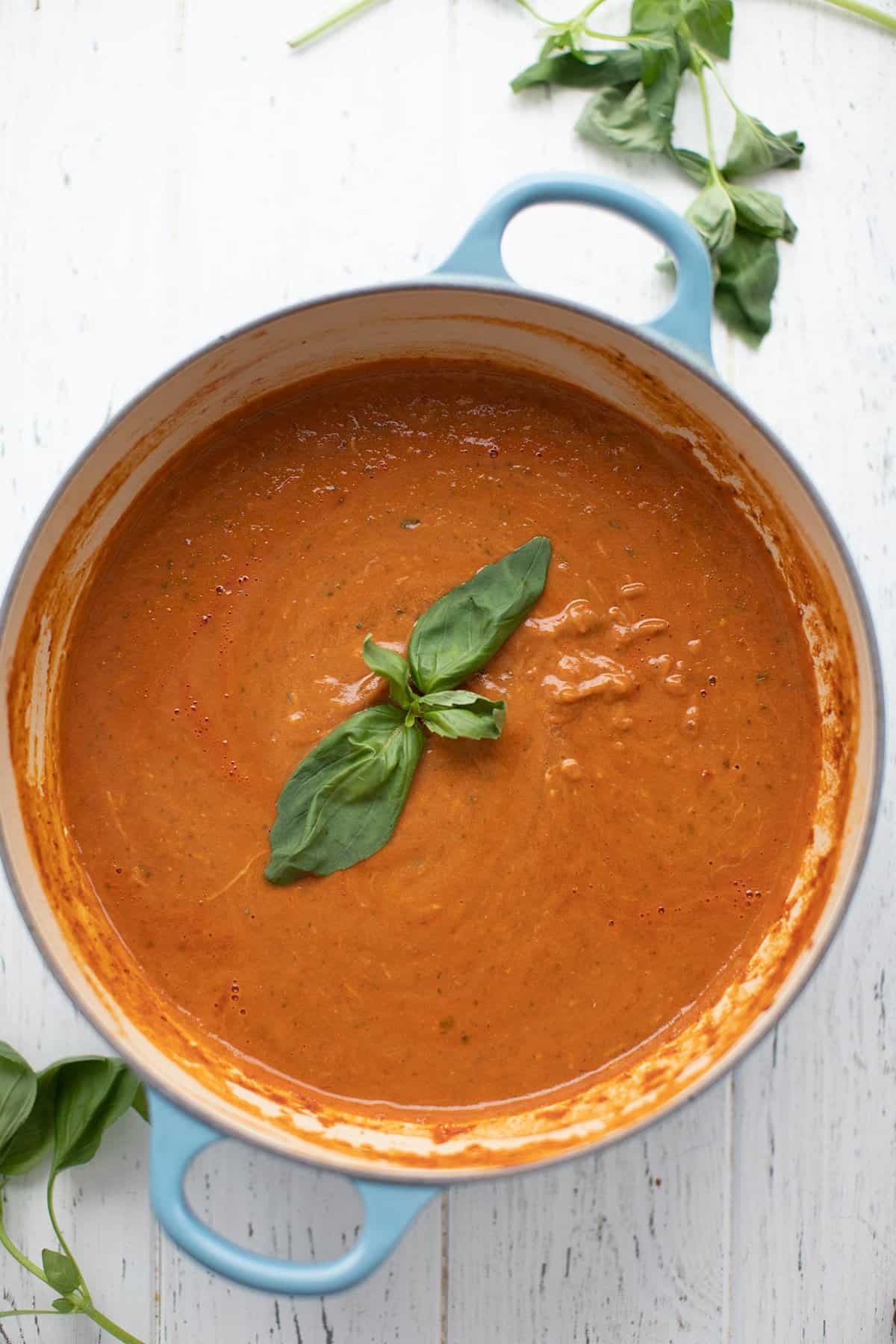 Top down image of a large blue dutch oven filled with keto tomato soup, with basil leaves floating on top. 