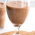 Titled image of a tall stemmed glass full of coffee protein shake on a small wooden cutting board.