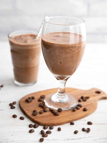 Two coffee protein shakes in glasses on a white wooden table with coffee beans strewn around.