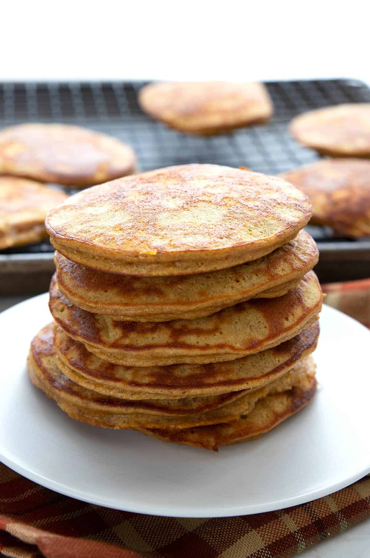 A stack of Keto Pumpkin Pancakes on a white plate, in front of a cooling rack with more pancakes.