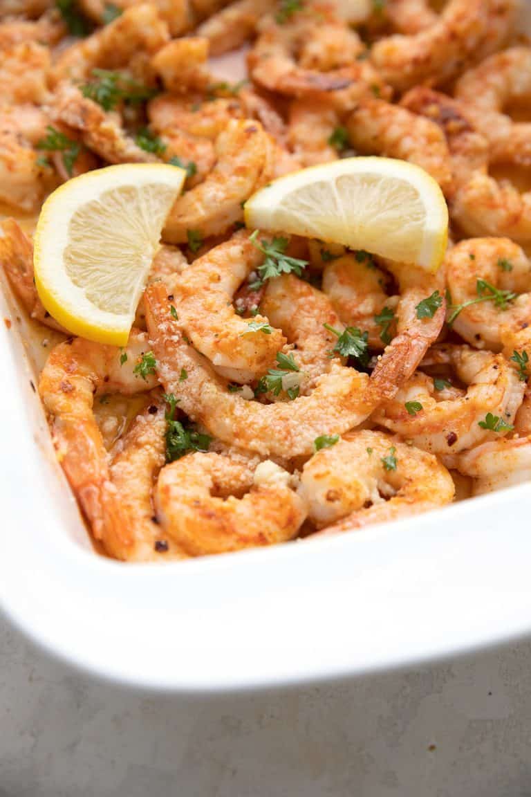 Close up shot of baked shrimp in a white baking dish, with lemon wedges.